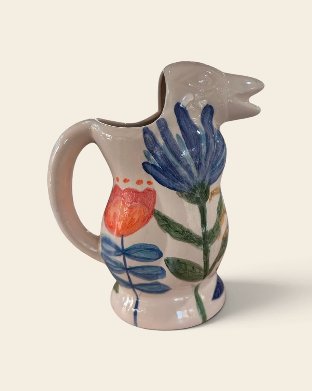 Jarra de cerámica color crema con el pico modelado como la cabeza de un pájaro, pintada a mano con una gran flor azul y un tulipán naranja de tallos verdes
