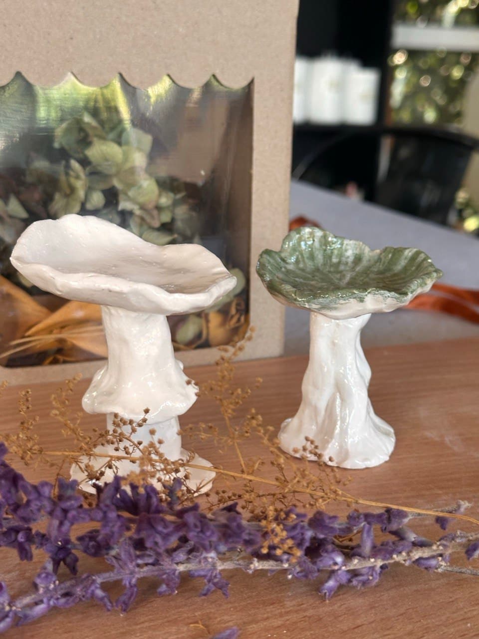 Dos hongos de cerámica modelados a mano sobre una mesa de madera, con ramitas de lavanda adelante y una caja de regalo de cartón con ventana atrás: uno todo blanco, de sombrero ondulado y con una pollerita en el tallo; el otro con el sombrero esmaltado verde salvia y el tallo blanco