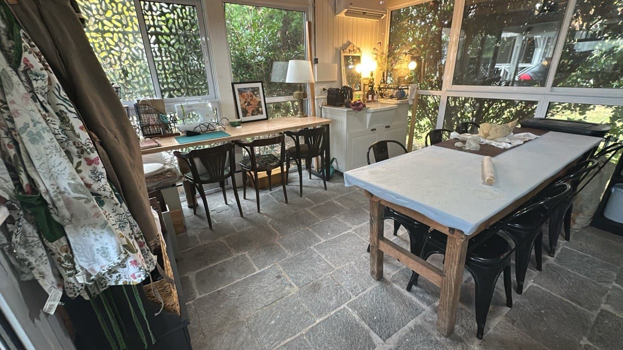 El taller de Cali en el jardín de invierno de su casa: una mesa larga de madera, sillas negras y grandes ventanales al jardín